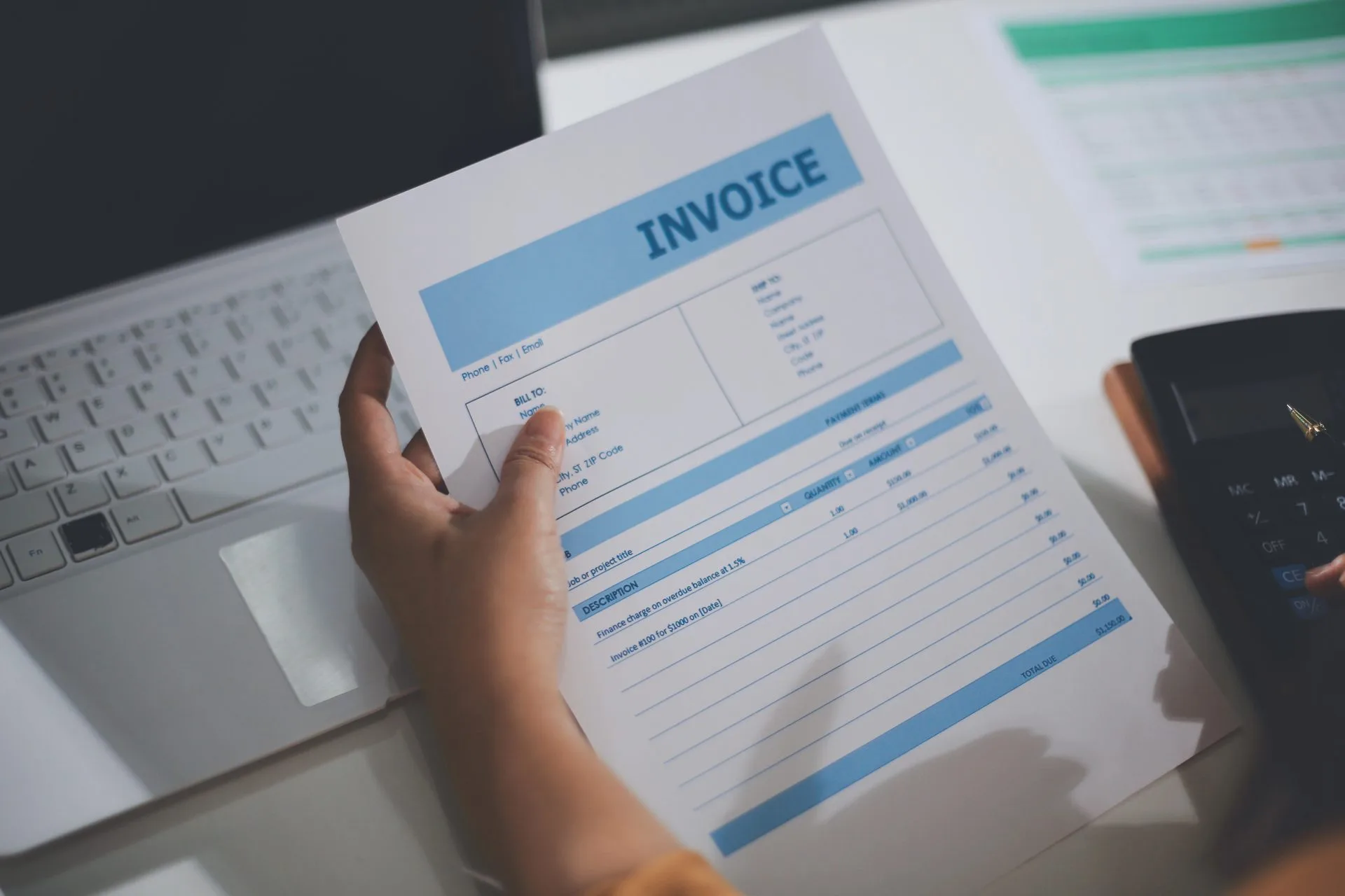 Person holding a printed invoice over a laptop and calculator at a desk
