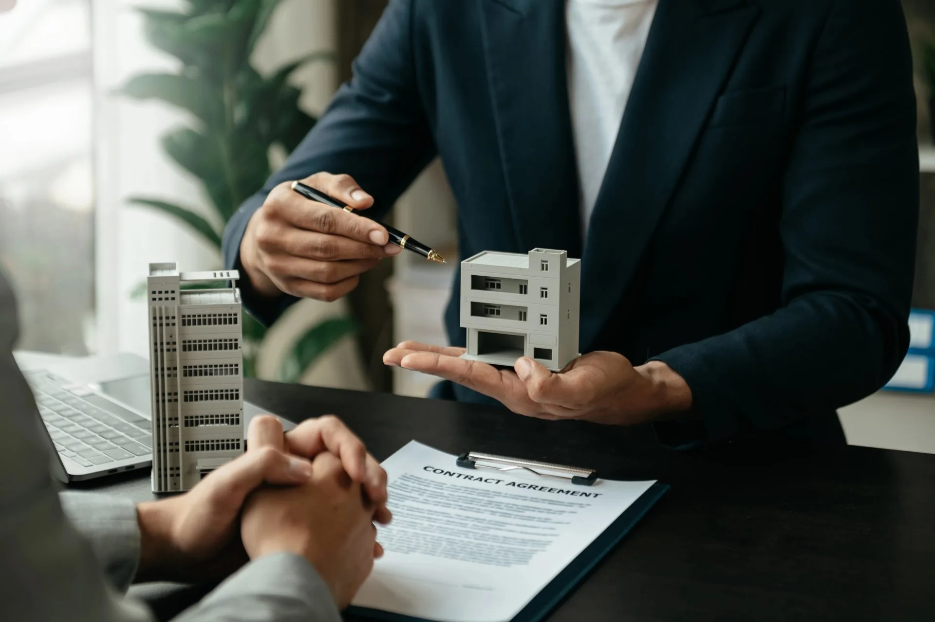 Man in a suit holding a model commercial building and pen while presenting a contract agreement across a desk