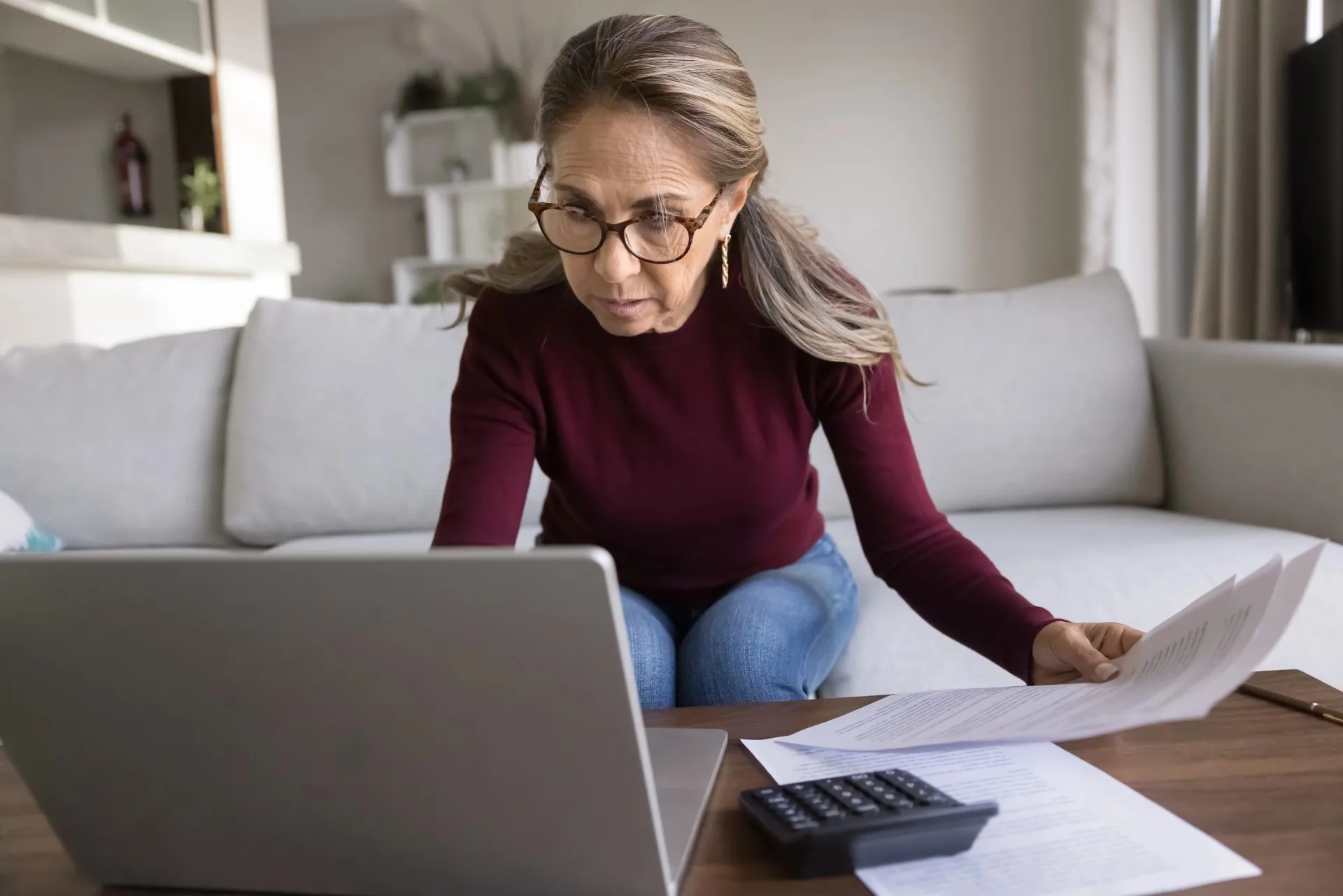 Woman in glasses reviewing loan documents at home with a laptop and calculator